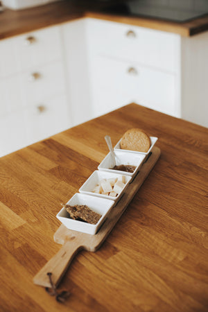 Smal serveringsbricka i trä med 4st vita små serveringsskålar fyllda med ost & kex stående på en köksbänk i trä