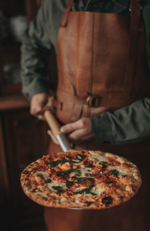 Man i läderförkläde hållandes pizzaspade med trähandtag & en pizza på med tomat, pesto & mozzarella som toppings 