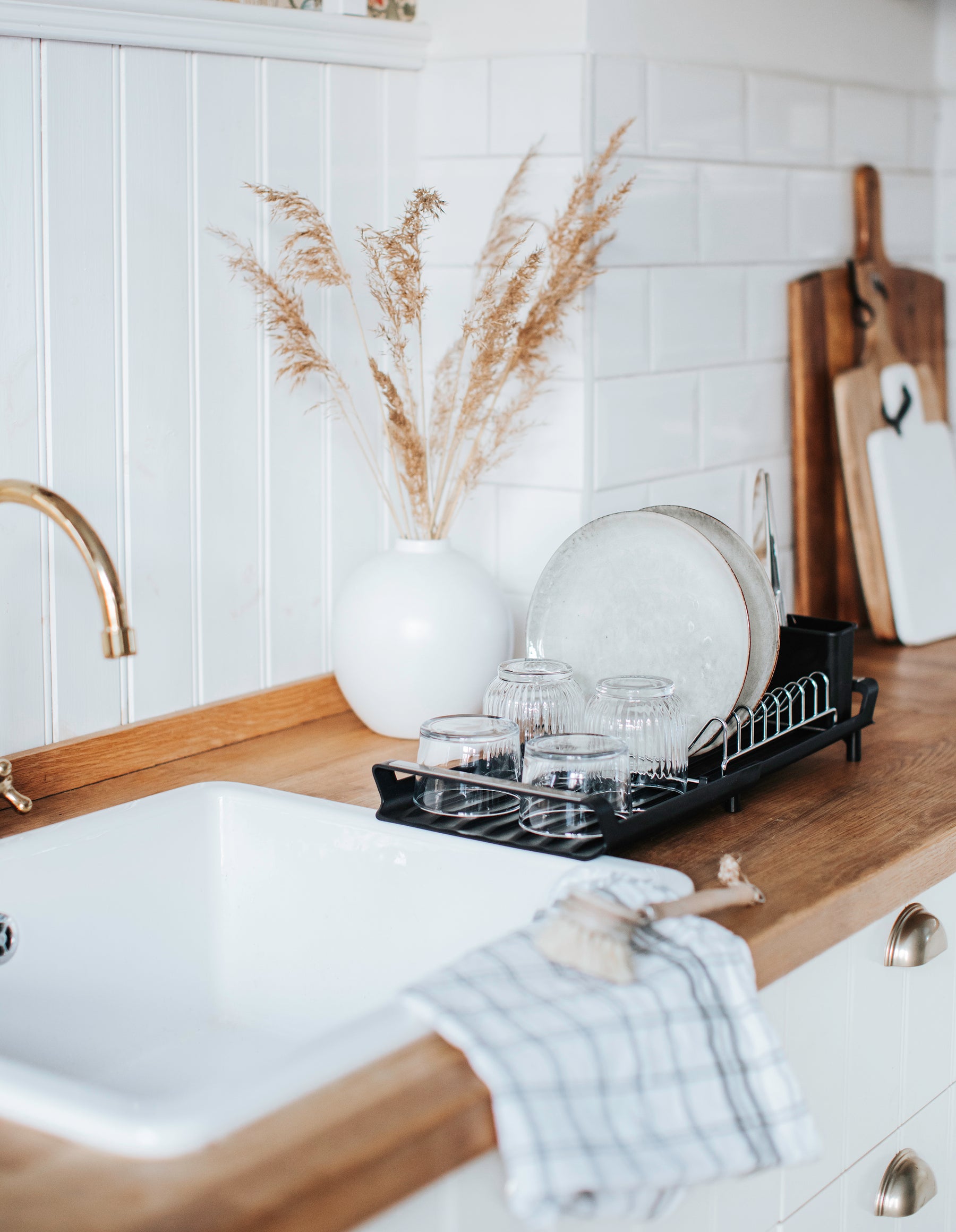 Vitt lantligt kök & vit vas med torkad vass placerad på köksbänk i trä. Dorre diskställ bredvid diskho med kopparkran